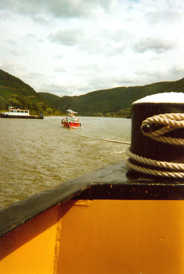 Kleines rotes Segelschiff hängt mit dickem Tau an einem Binnenschiffpoller, ruhige Mosel, Berge.