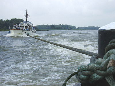 Weißes Sportboot hängt mit dickem Tau an einem Binnenschiffpoller, in aufgewühltem Rheinwasser.